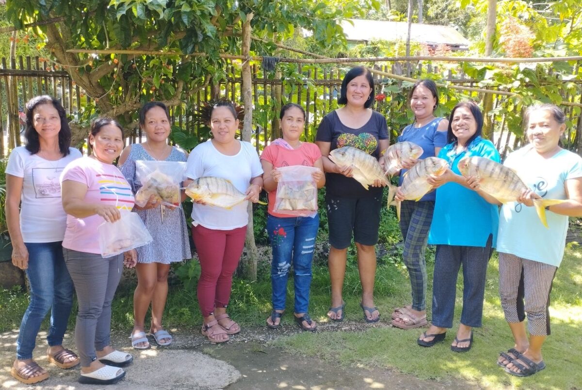 Nine women from the Bogtong Fish Processors association stand in a row, smiling and holding up fish and fish products