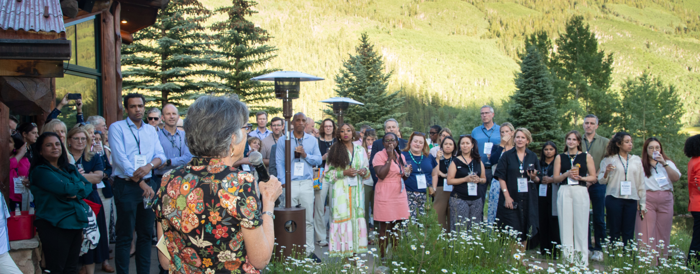 A group of attendees at the Aspen Business and Society Summit, standing in a circle on a green lawn, with green fir trees and a mountain bathed in sunlight behind them