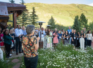 A group of attendees at the Aspen Business and Society Summit, standing in a circle on a green lawn, with green fir trees and a mountain bathed in sunlight behind them