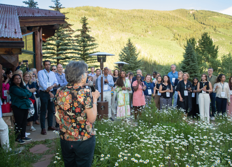 A group of attendees at the Aspen Business and Society Summit, standing in a circle on a green lawn, with green fir trees and a mountain bathed in sunlight behind them