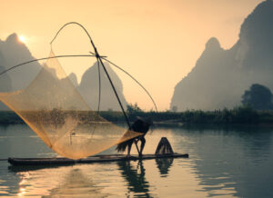 Sunrise over water, with mountains in the background and a fisherman with a net on a raft in the foreground