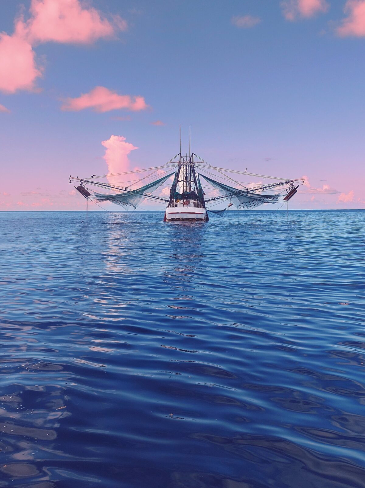 A fishing boat photographed head on with its nets stretched out to the side and a broad expanse of blue water under it and a pink and blue sunset above