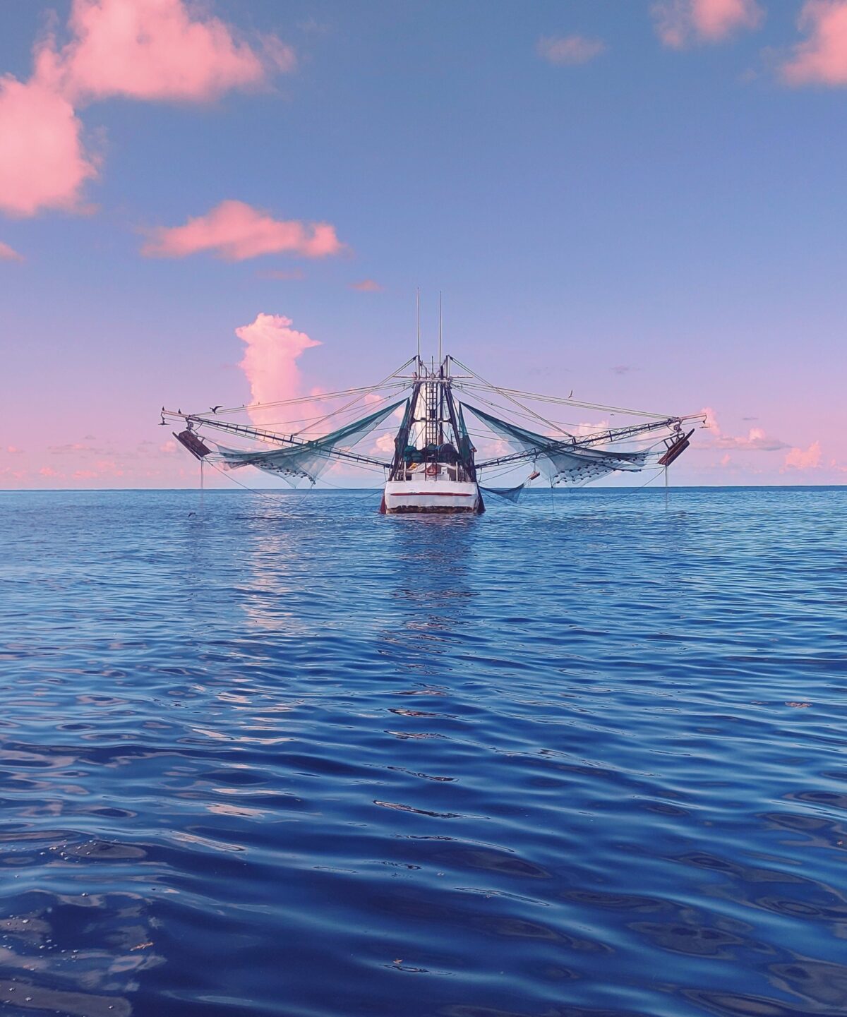 A fishing boat photographed head on with its nets stretched out to the side and a broad expanse of blue water under it and a pink and blue sunset above