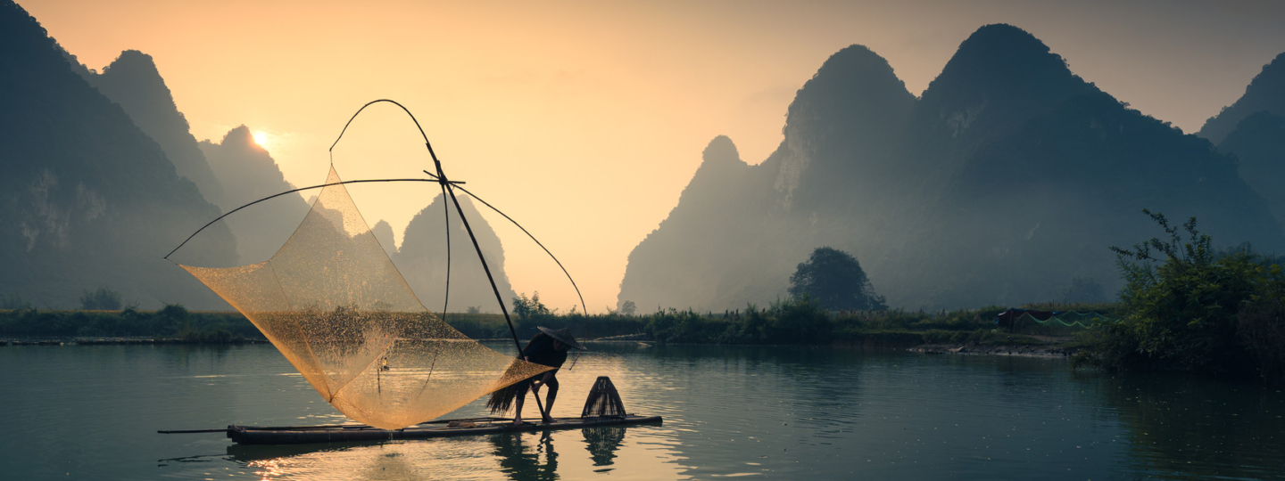 Sunrise over water, with mountains in the background and a fisherman with a net on a raft in the foreground