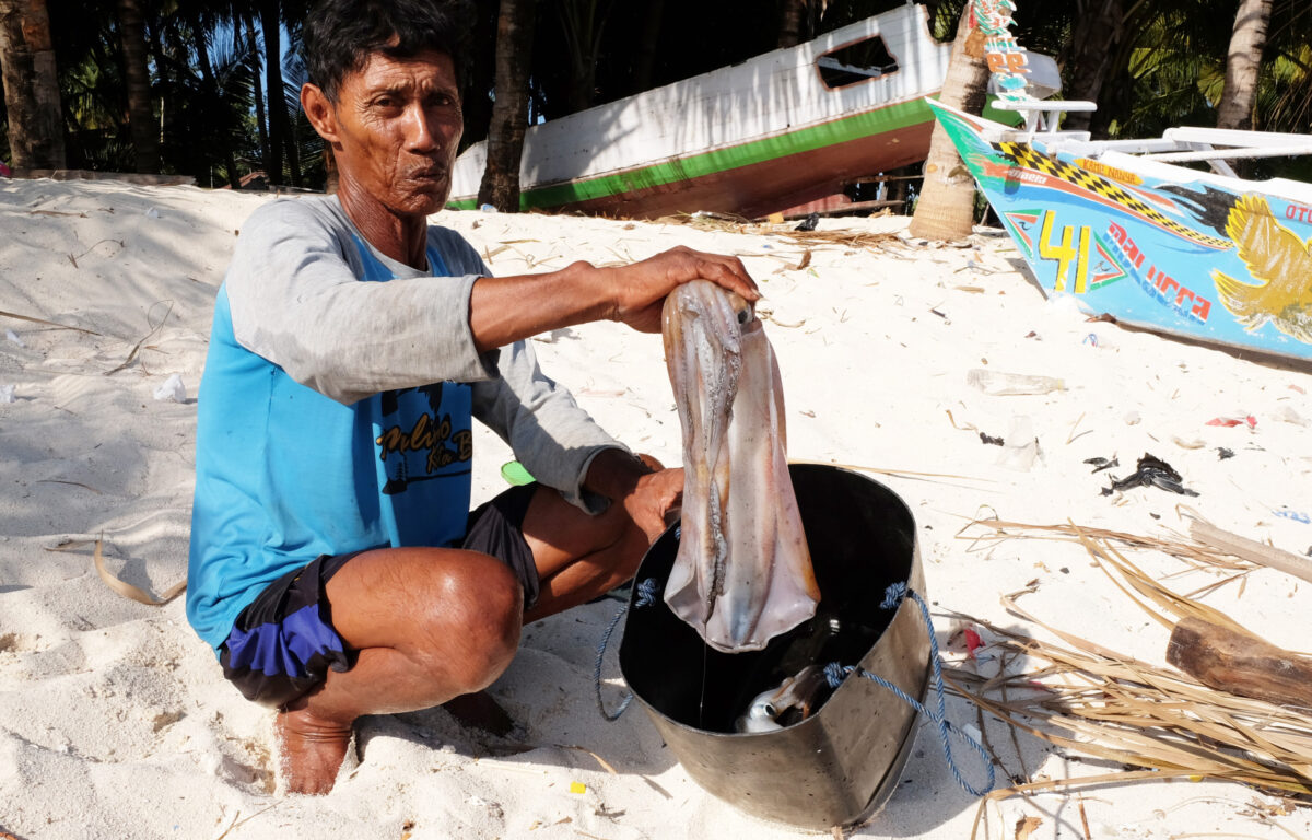 A man crouched on the sand, holding a freshly caught octopus over a metal pot