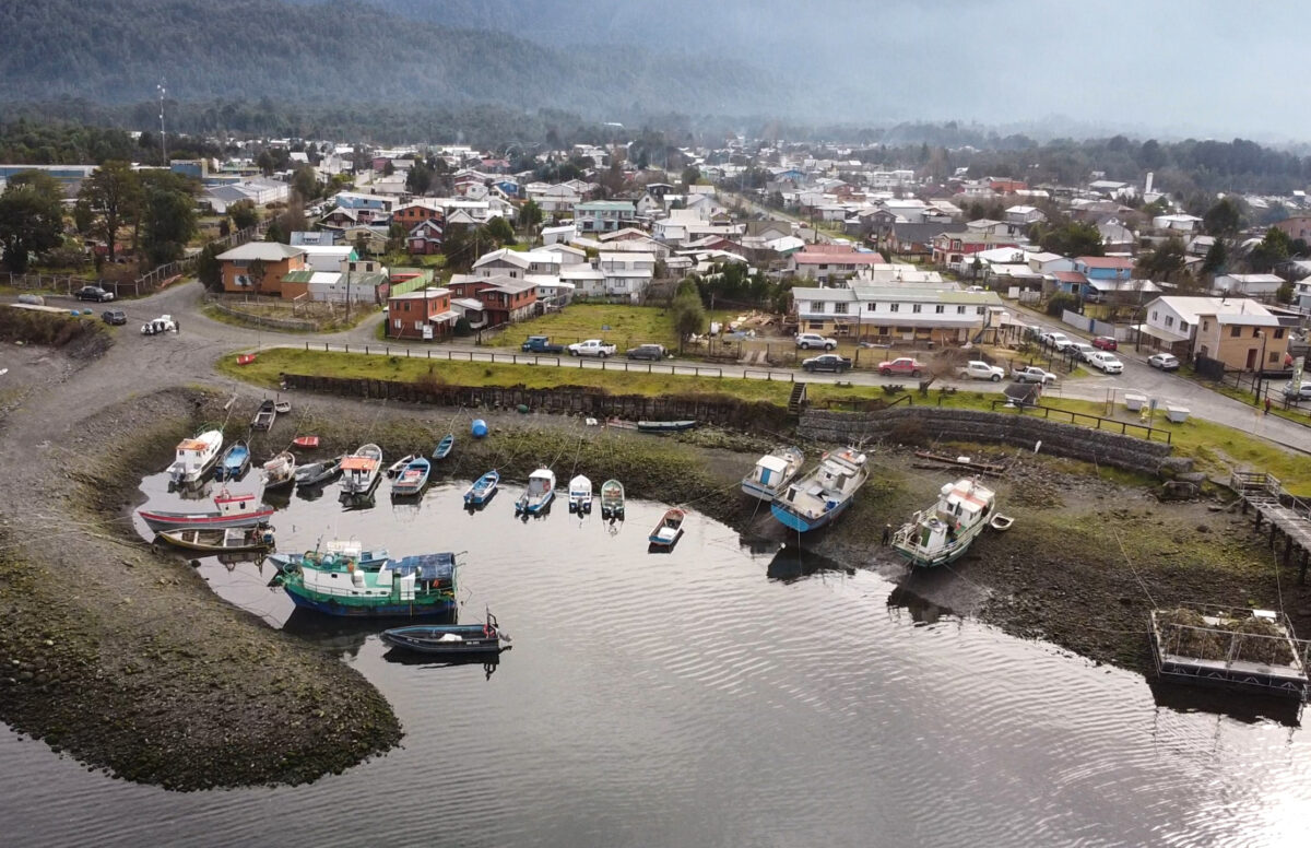 An aerial view of a town in southern Chile, with many buildings close to the shore and fishing boats lined up in a protected cove