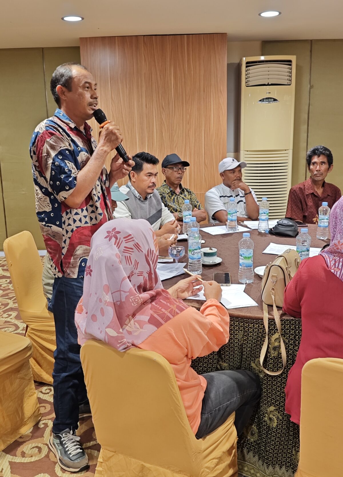 A man standing with a microphone addressing a group of people sitting around a table