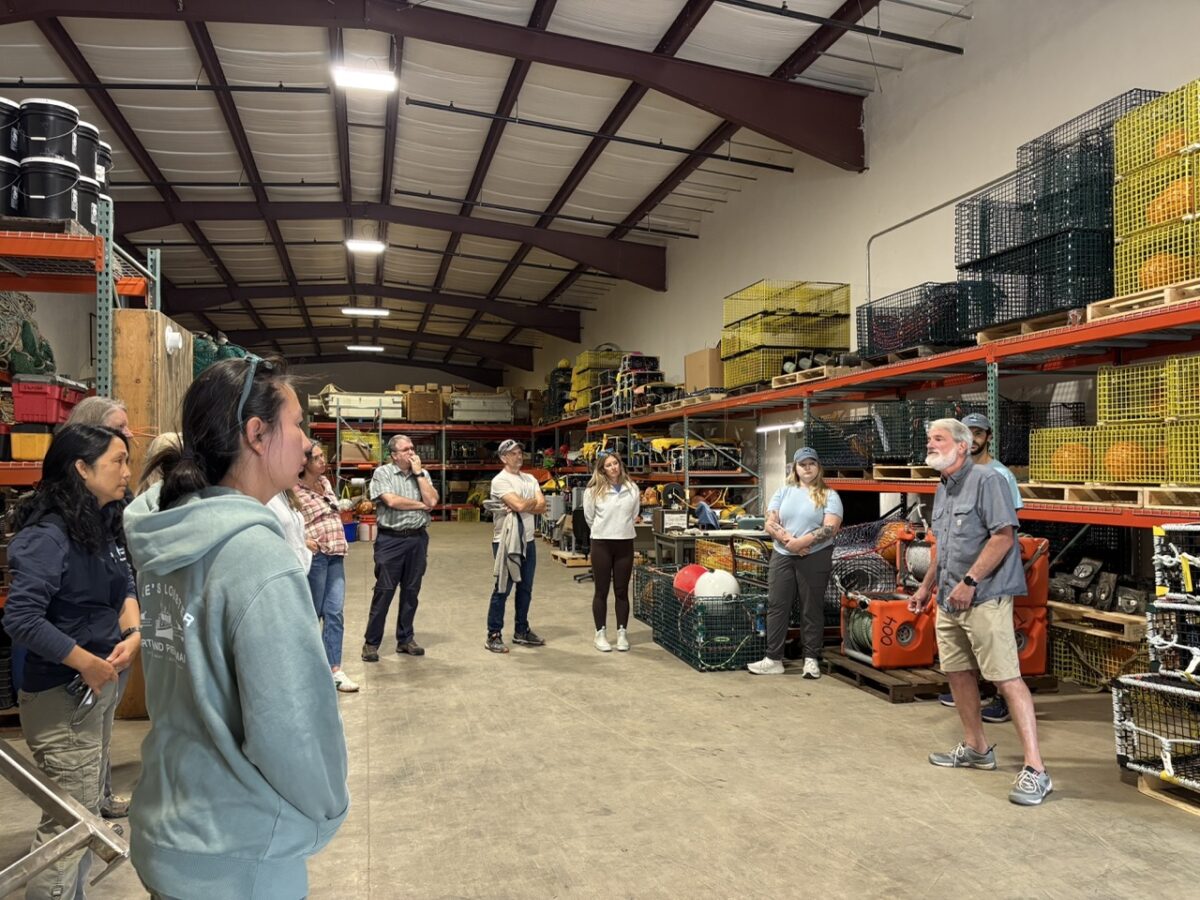 A group of people standing in a large circle listening to presentation about on-demand gear, inside a large warehouse with shelves stacked with gear
