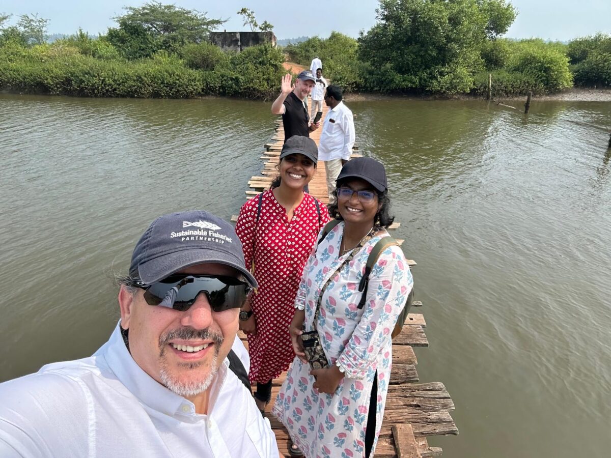 SFP CEO Jim Cannon, in a baseball cap and sunglasses, in front of a group of people walking on a bridge over shrimp farms by mangroves