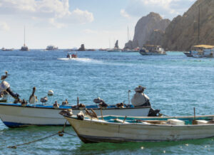 Three small fishing boats lined up in a bay in Cabo San Lucas, Mexico, with a rocky, hilly coastline behind them and white fluffy clouds above