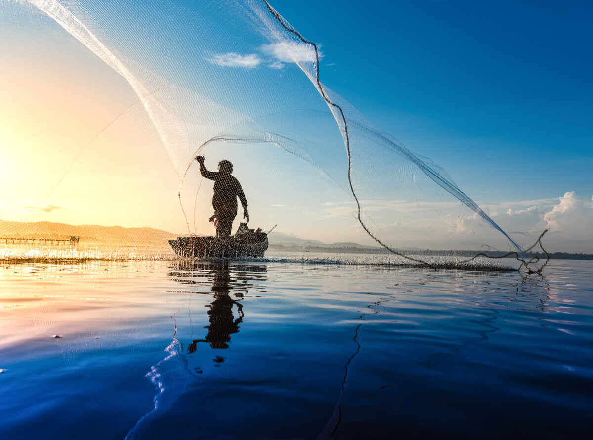 A fisherman standing on a boat in the middle of a lake throwing a net, with a yellow sunrise behind him