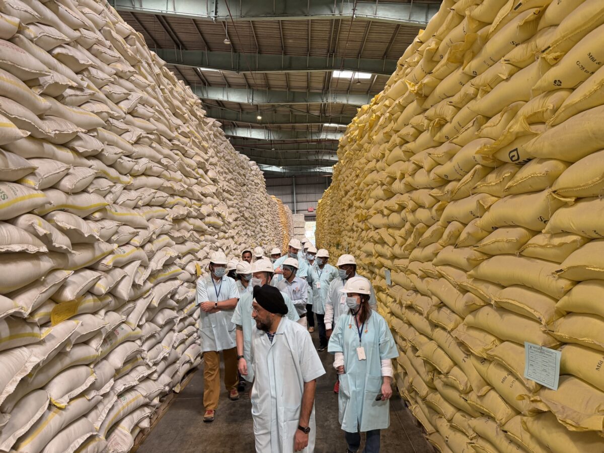 A group of people dressed in protective smocks and hats, standing in an aisle between two enormous stacks of bagged fish feed