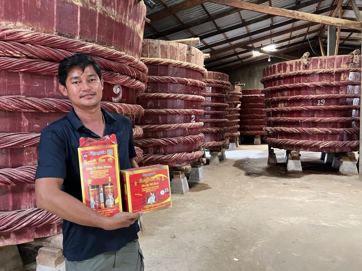 A man holding packages of fish sauce and standing in front of a row of traditional vats for making fish sauce at a producer facility.