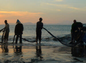 Fishermen bringing in nets onto a fishing boat on the beach at sunset