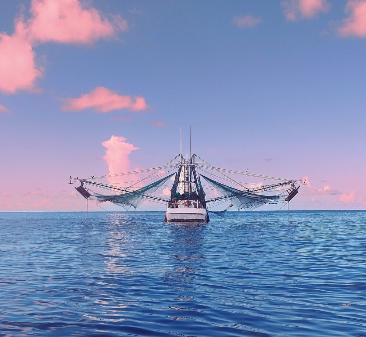 Fishing boat in the Gulf of Mexico with its trawls raised, against a pink and blue sunset